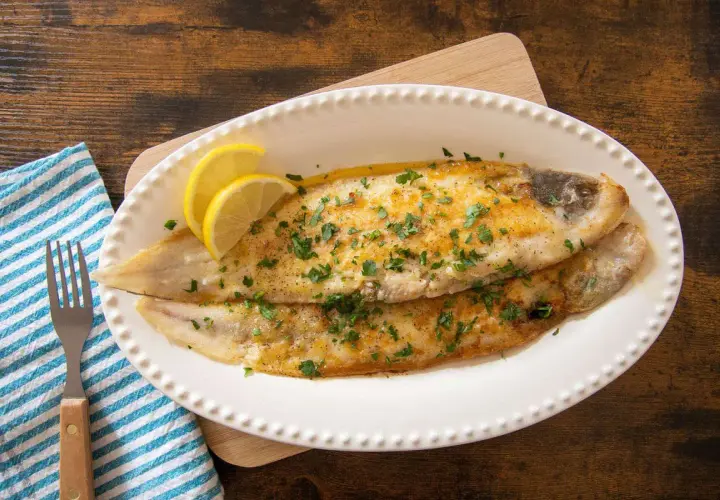 Due filetti di sogliola alla parmigiana dorati, guarniti con prezzemolo tritato e spicchi di limone, serviti su un piatto bianco.