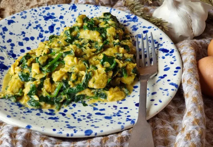 Un delicioso revuelto de espinacas cremoso, servido en un plato con salpicaduras azules. Acompañado de pan, ajo y huevos frescos.