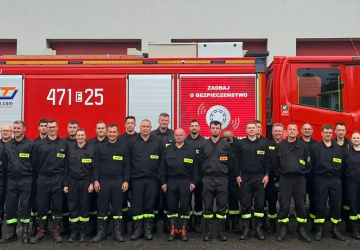 Grupa strażaków w mundurach, obok czerwonego wozu strażackiego. Dowódca sekcji OSP stoi w pierwszym rzędzie, w jasnym mundurze.