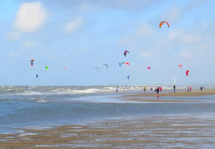 Plaża z wieloma latawcami do kitesurfingu na niebie i ludźmi spacerującymi po piasku.