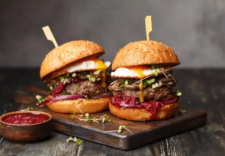 Dwa soczyste burgery z jajkiem sadzonym i dodatkami na desce, obok miseczka sosu.
