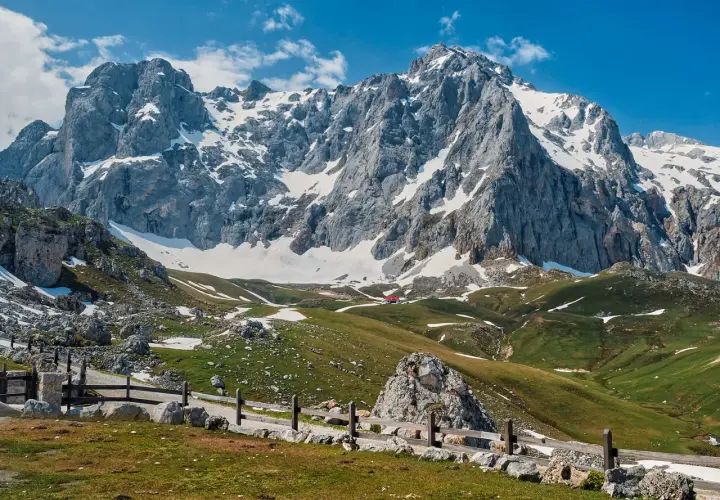 Majestuosas montañas nevadas y verdes praderas invitan al senderismo en los Picos de Europa. Un refugio rojo se asoma entre las colinas.