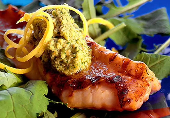 Grillad havsabborre med en grön topping och citronskal, serverad på en bädd av gröna blad.
