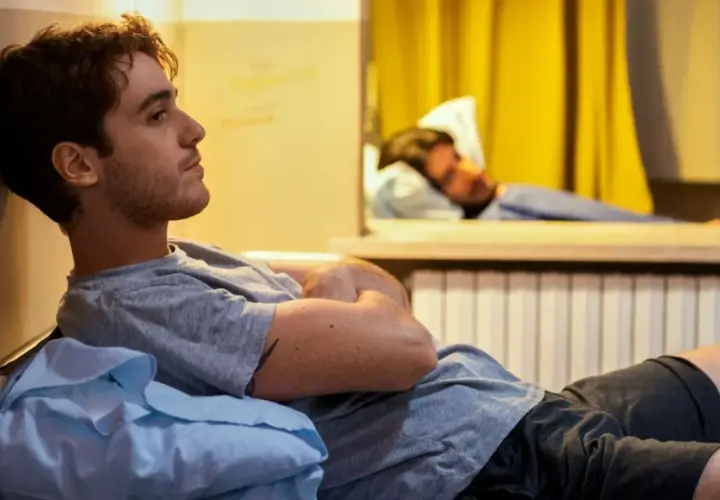 Un giovane uomo pensieroso, forse un genio incompreso o in preda alla follia, riposa su un letto in una stanza con tende gialle.