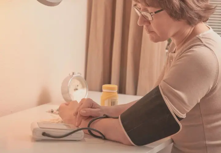 Kobieta z polsartem mierzy ciśnienie. Na stole leżą tabletki i budzik.