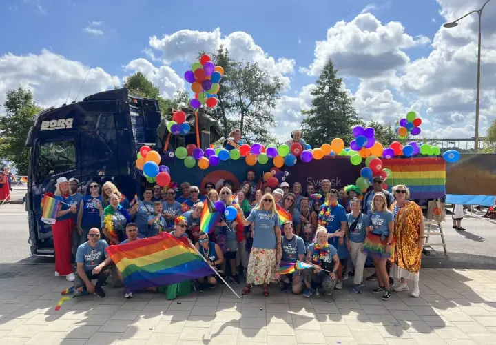 Stor grupp människor firar med regnbågsflaggor och ballonger vid en pride park.