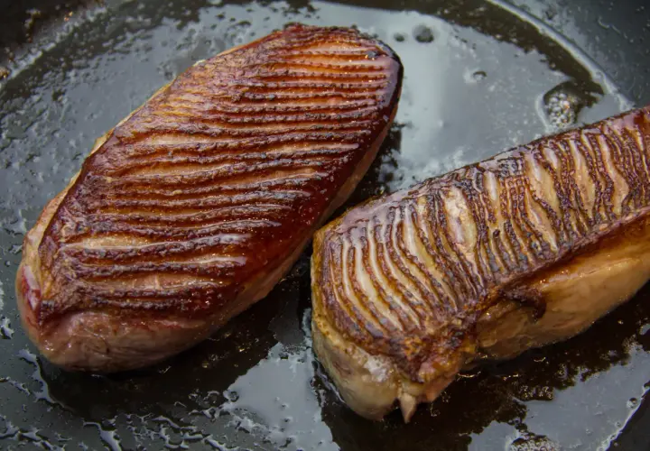 Deux magrets de canard grillés dans une poêle, prêts pour une recette savoureuse. La peau est dorée et croustillante.