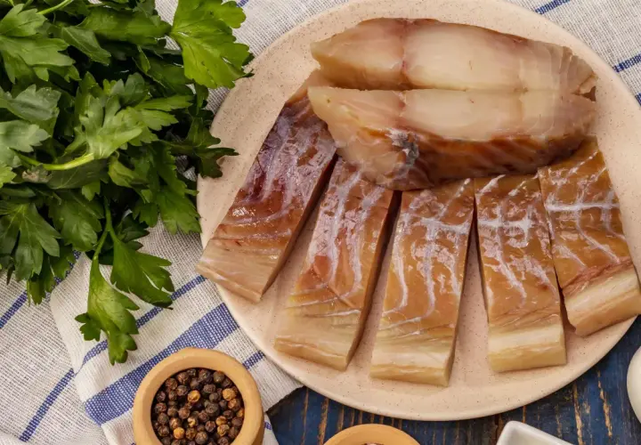 Kawałki wędzonego dorsza czarnego na talerzu, obok pęczek natki pietruszki i miseczka z pieprzem.