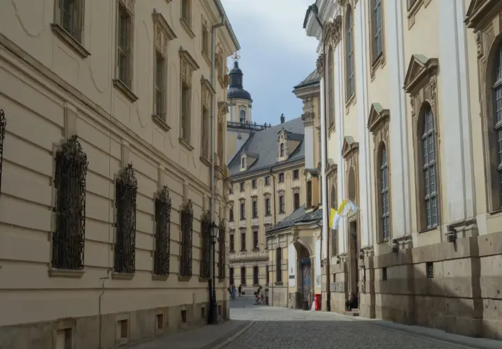 Wąska, brukowana uliczka prowadzi do serca starego rynku we Wrocławiu. Po bokach piętrzą się zabytkowe kamienice z ozdobnymi fasadami i kratami w oknach.