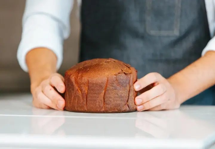 Manos sostienen un pan brioche dorado y esponjoso sobre una superficie blanca.