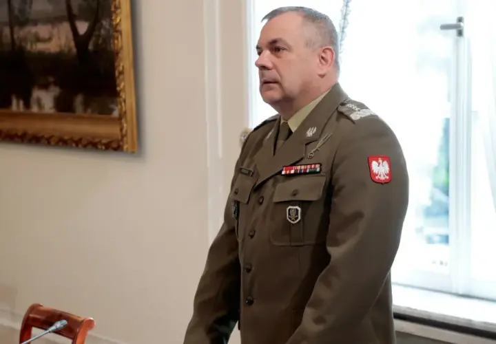 Generał w mundurze, z orłem na piersi, stoi w sali, gdzie odbywa się spotkanie korpusu generałów.