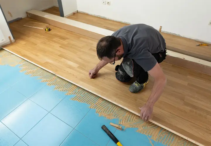 Pose d'un parquet stratifié sur un sol bleu carrelé, avec de la colle appliquée. L'homme ajuste une lame de bois.