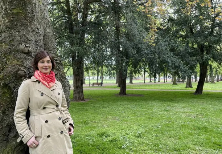 Kristina Hartenbach lehnt an einem Baum im Park. Sie trägt einen beigen Trenchcoat und einen roten Schal.