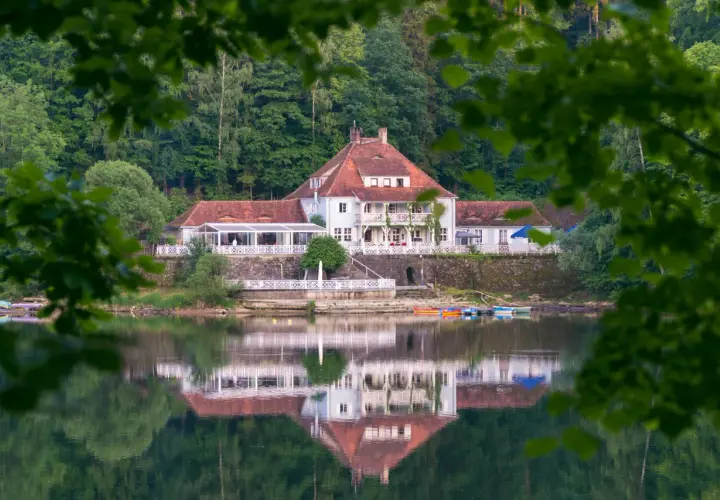 Biały hotel z czerwonym dachem nad spokojnym jeziorem, odbijający się w wodzie.