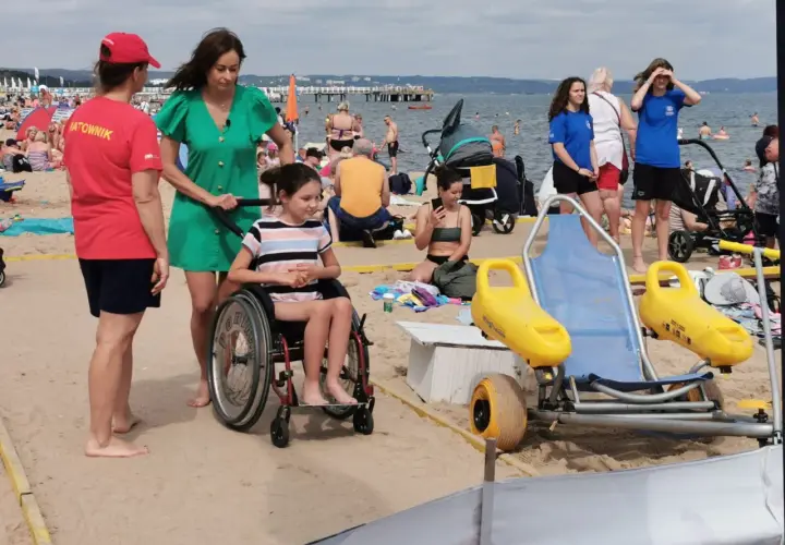 Na plaży dla niepełnosprawnych ratowniczka pomaga dziewczynce na wózku inwalidzkim. W tle widać innych plażowiczów i morze.