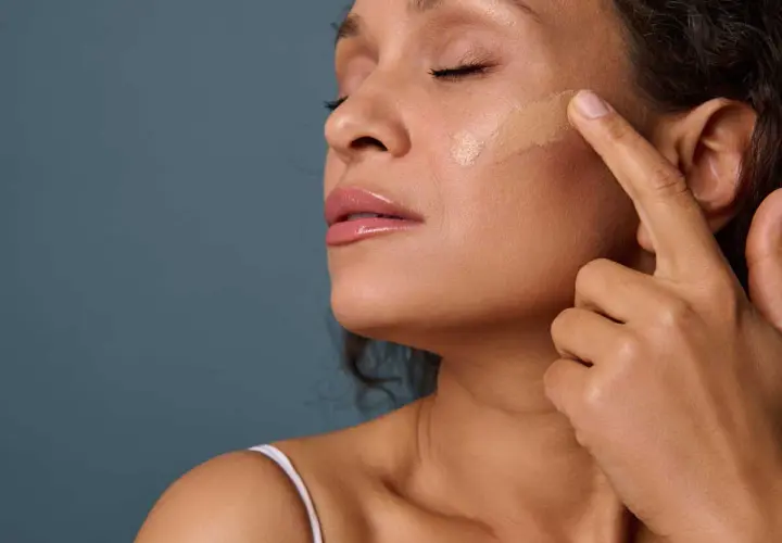 Application d'un fond de teint pour un maquillage teint parfait. Une femme applique délicatement le produit sur sa joue.