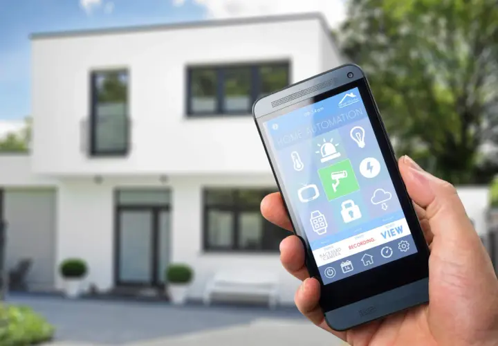 Dłoń trzyma smartfon z aplikacją do **instalacji inteligentnego domu**. Na ekranie widoczne ikony sterowania oświetleniem, ogrzewaniem, kamerami i innymi funkcjami.