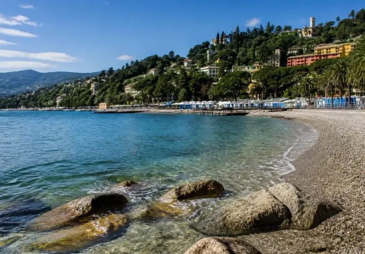 Plage de galets à Rapallo, avec des cabines bleues et blanches alignées, des maisons colorées sur la colline verdoyante et une mer bleue étincelante.