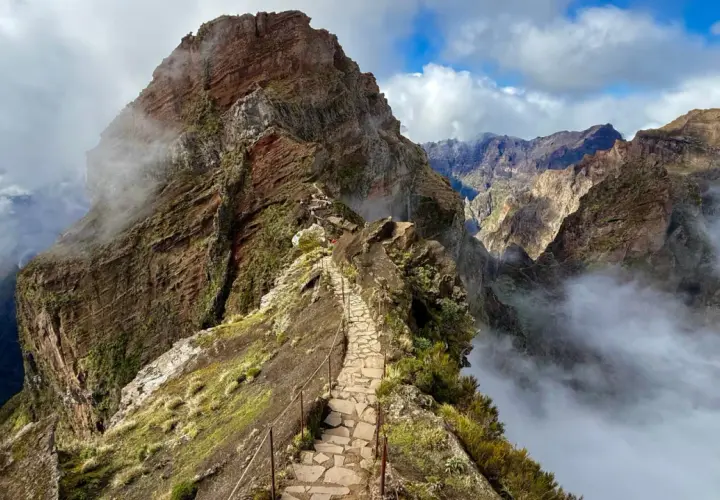 A stone path winds along a narrow mountain ridge, offering what is likely the best hiking in Portugal. Clouds drift through the dramatic, rugged peaks.