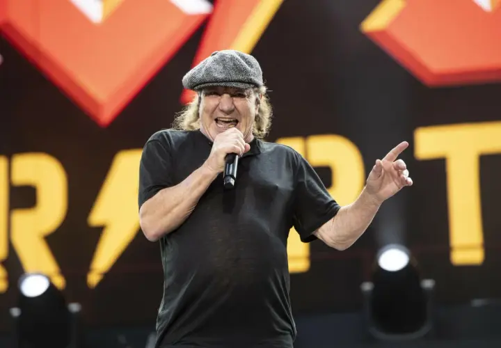 Brian Johnson young, w czapce i czarnej koszulce, śpiewa do mikrofonu na tle sceny z logo zespołu.