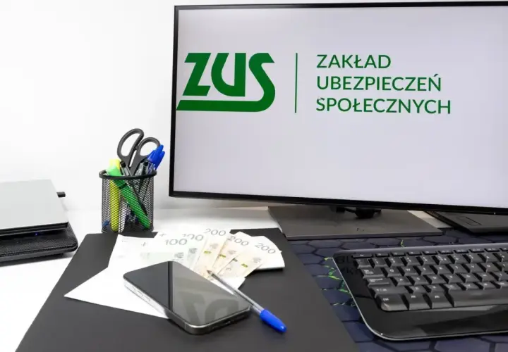 Na biurku leżą pieniądze, telefon i długopis. Na monitorze widnieje logo ZUS, symbolizujące składkę ZUS.