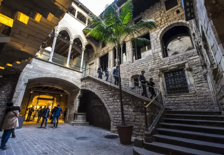 Un patio interior de piedra en el Born, con una palmera y escaleras que suben. Gente pasea por el lugar.