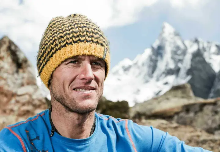 Stephan Siegrist, ein Bergsteiger, lächelt in die Kamera, eine gestreifte Mütze auf dem Kopf, vor einer schneebedeckten Bergkulisse.