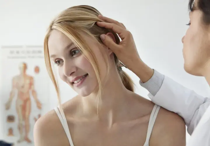 Dermatolog bada skórę głowy młodej kobiety. W tle wisi plakat anatomiczny.