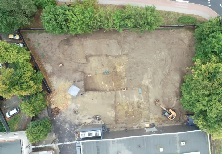 Wykop archeologiczny na placu budowy, gdzie prowadzone są badania przed pozwoleniem na budowę. Widoczny koparka i ślady dawnej zabudowy.
