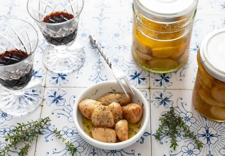 Ail confit au four, tendre et parfumé, servi dans un bol avec des herbes. Deux verres de vin rouge et des bocaux d'ail confit complètent la scène.