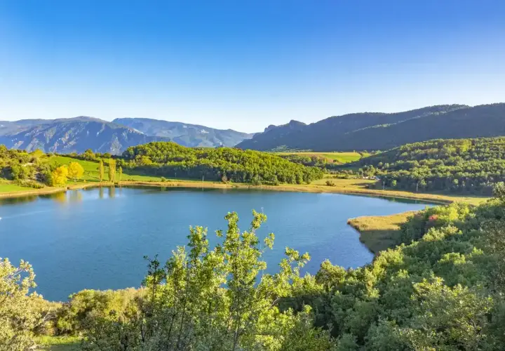 Paisaje sereno en la conca de dalt: un lago azul rodeado de colinas verdes y montañas bajo un cielo despejado.