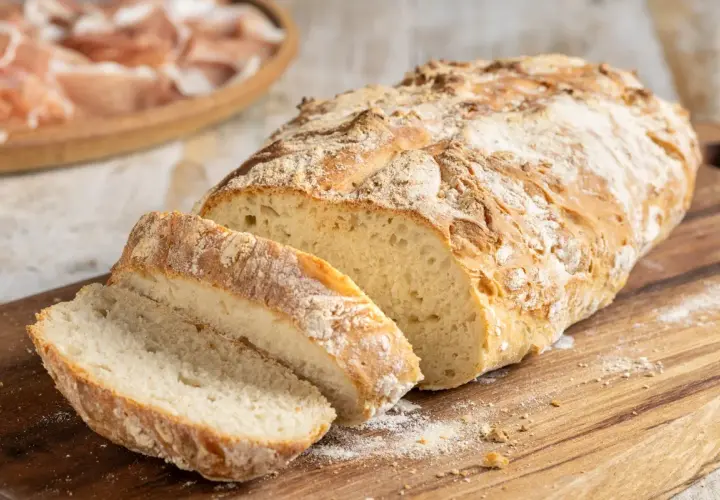 Pane fatto in casa, croccante fuori e soffice dentro, tagliato su un tagliere di legno. Sullo sfondo, un piatto di affettati.