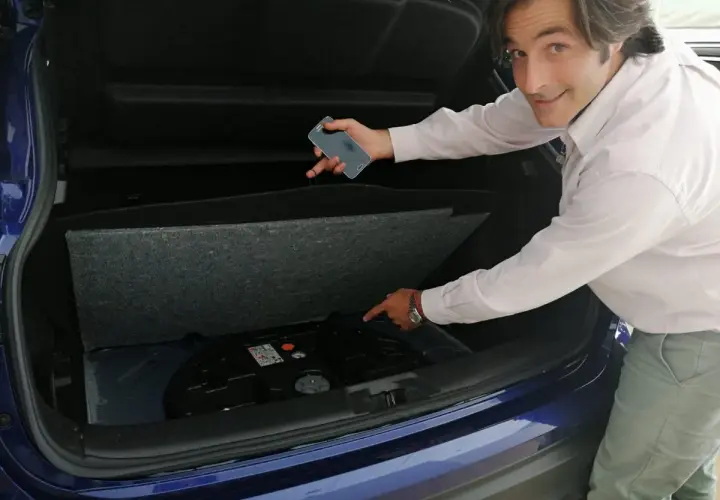 Hombre señala el espacio vacío en el maletero de un coche azul, donde debería estar la rueda de repuesto. "He pinchado y no tengo rueda de repuesto", parece decir.