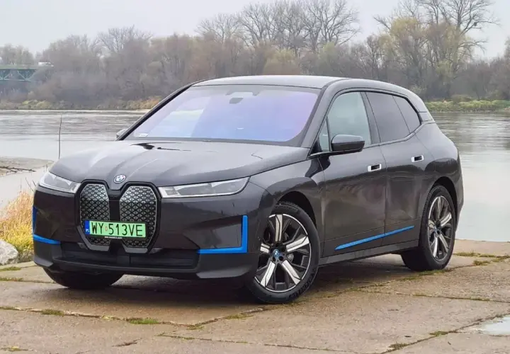 Ciemnoszary BMW iX z niebieskimi akcentami stoi nad rzeką, z mostem w tle.