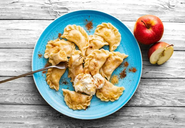Pyszne pierogi z jabłkami, posypane cynamonem, na niebieskim talerzu, obok jabłka.
