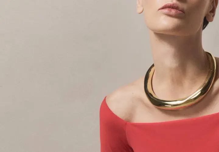 A woman wears a bold gold necklace with a curved neckline, complementing her red off-the-shoulder top.