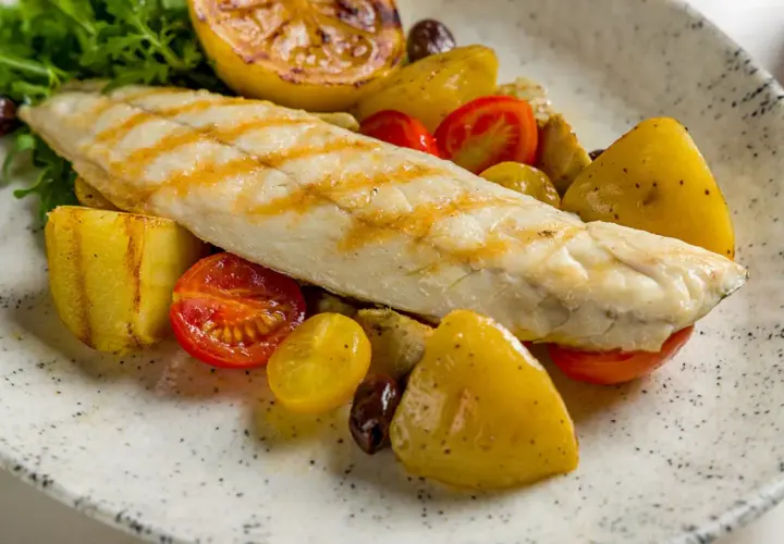 Filetto di persico grigliato su un piatto con patate arrosto, pomodorini, olive e limone caramellato.