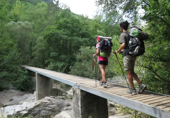 Dos excursionistas con mochilas cruzan un puente de madera en una ruta transpirenaica.