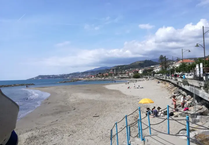 Plage de sable à Santo Stefano al Mare, avec des gens profitant du soleil et de la mer. La ville s'étend le long de la côte, bordée de collines verdoyantes.
