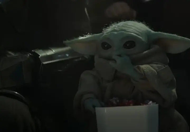 Dziecko z mandalorian film, w brązowym płaszczu, je słodycze z białego pudełka. Obok widać fragment zbroi Mandalorianina.