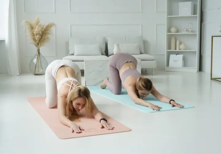 Dwie kobiety ćwiczą jogę ashtanga w pozycji dziecka, rozciągając się na matach w jasnym, minimalistycznym wnętrzu.