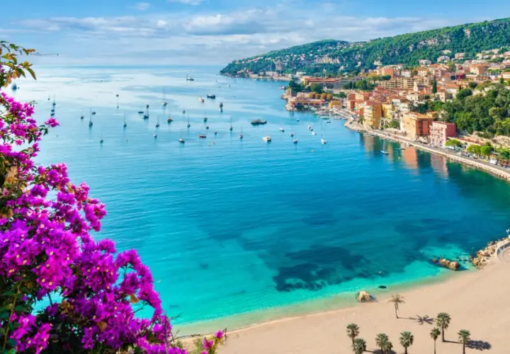 Blick auf einen malerischen Strand in Frankreich mit türkisfarbenem Wasser, Segelbooten und einer Küstenstadt, umrahmt von leuchtend lila Bougainvillea.