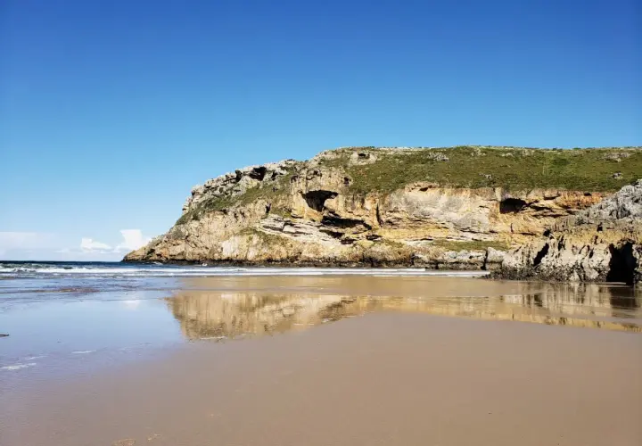 Reflejos dorados en la arena mojada de la playa de Fuentes, con acantilados cubiertos de verde bajo un cielo azul.