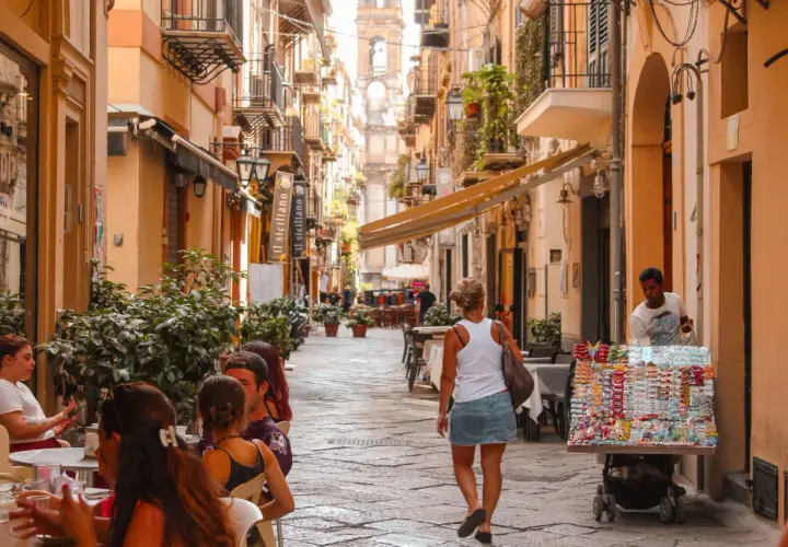 A woman walks down a bustling street in Sicily, past shops and cafes. The scene suggests a vibrant, lively atmosphere, answering the question: is Sicily safe for tourists to enjoy?