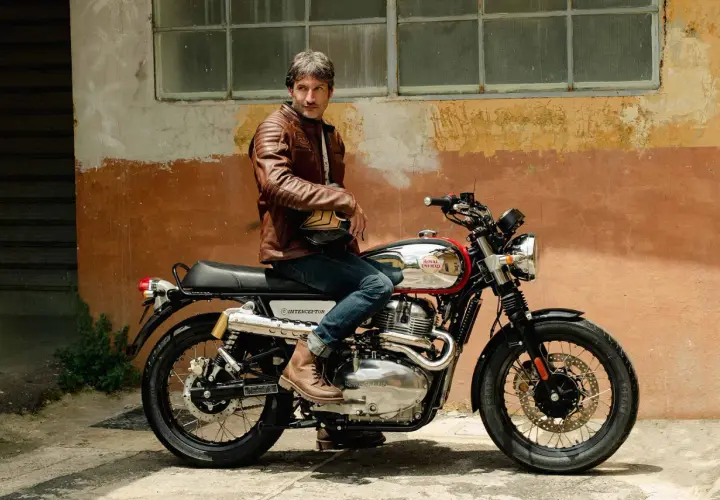 Uomo con giacca in pelle marrone seduto su una moto Royal Enfield Interceptor. I terminali moto cromati brillano sotto il sole.