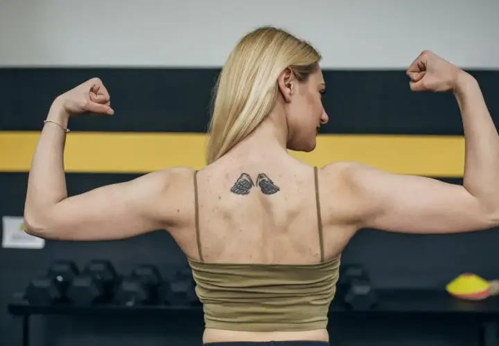 Silna kobieta z tatuażem skrzydeł na plecach napina biceps. Zastanawia się, ile po tatuażu nie można ćwiczyć, by go nie uszkodzić.