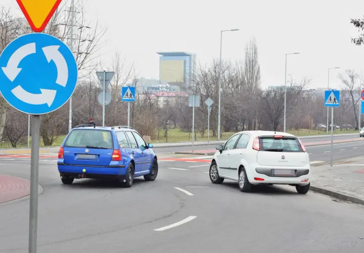 Niebieski Volkswagen i biały Fiat na rondzie. Kierowcy zastanawiają się, który zjazd z ronda na który pas wybrać, by bezpiecznie kontynuować jazdę.