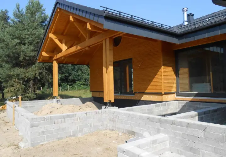 Drewniany dach krokwiowy nad wejściem do domu w budowie, z widocznymi belkami i elementami konstrukcyjnymi.