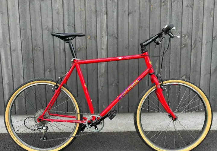A vintage red Gary Fisher Advance mountain bike with tan tires stands against a wooden fence.