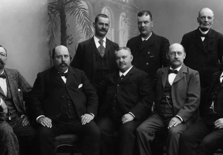 Gruppbild på män i 1900 talet kläder. De bär kostymer och flugor, redo för ett formellt porträtt.